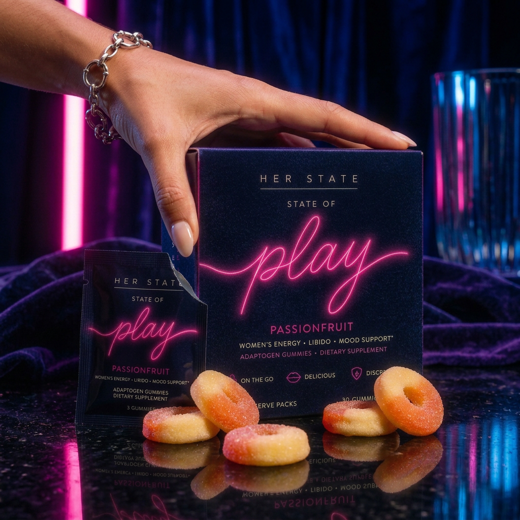 A hand resting on the navy velvet Her State box, its neon pink play script glowing, with passionfruit gummy rings in front